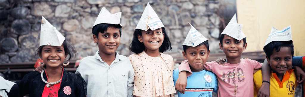 A line of cheerful Indian children smiling and wearing paper hats they crafted during a workshop
