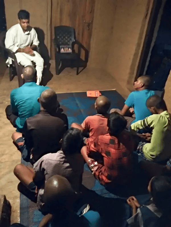 A community volunteer addressing rural residents gathered indoors during a public awareness and neighborhood dialogue session